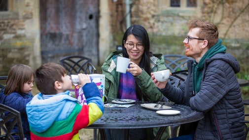A family of two adults and two young children eating outside the Courtyard Cafe at Lacock, they're wearing winter coats and smiling with mugs in their hands.